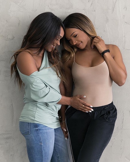 Khadijah touching Malika's belly as they gush and pose standing up against the wall.