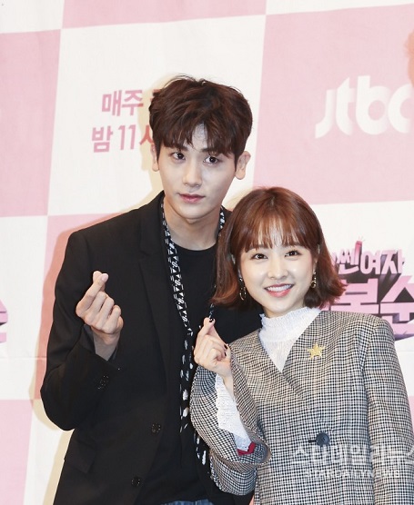 Bo-Young leaning in front of Hyung-Sik at the press conference of 'Strong Girl Bong-Soon', both showing the Korean Love symbol with their hands.