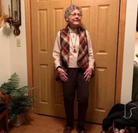 Patricia's picture where she is standing near a bedroom door clad in a sweater inside a jacket.