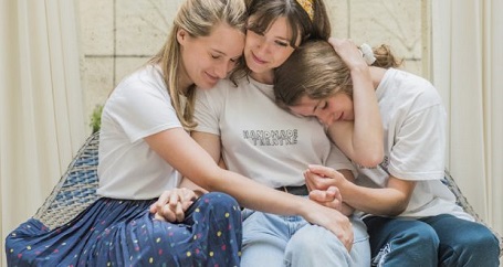 Three Girls sitting on the end-side of a bed as they bundle up together feeling sad and fulfilled at the same time. They are the cast of 'Di and Viv and Rose'.