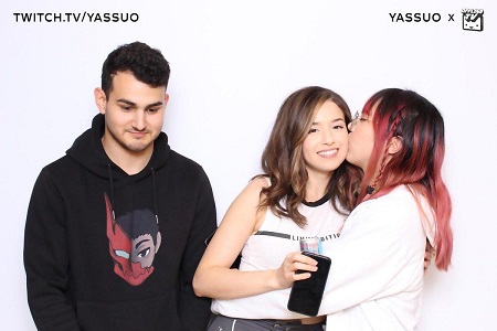 Hasaan in a black hoodie looks disconcerted as the Korean Lily in red-flame hair gives a kiss on the cheek to Pokimane while hugging her and Poki is holding her camera in her right hand as she looks at the camera.