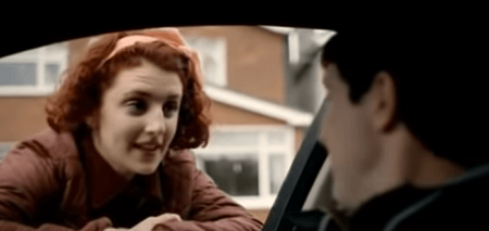 A scene from Love/Hate as Kayleigh peeks into a car window resting her hands on the window with her red hair as the guy in the car is blurry.