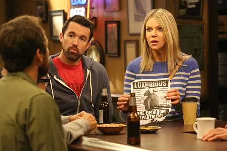 A scene from the show, Dee holds a B\W ad-post as Mac looks in confident in his grey jacket.