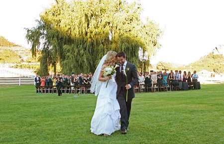 The couple smiling and walking together after their wedding as the guests all stand up in the background to look at them.