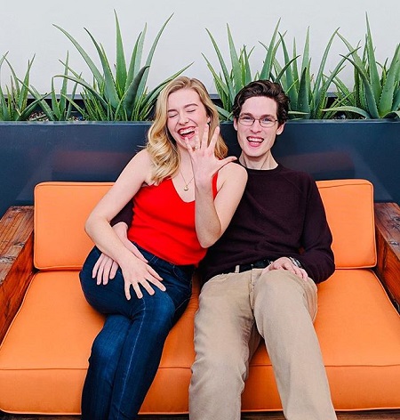 McMann and Sam sitting on a couch, McMann showing-off her ring by raising her left hand, fingers spread.