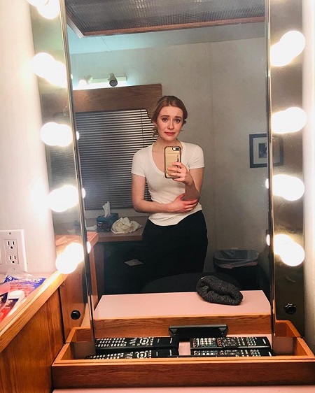 A far mirror selfie by McMann in white T shirt looking a little pale and holding her right hand (with the phone) with her left hand.