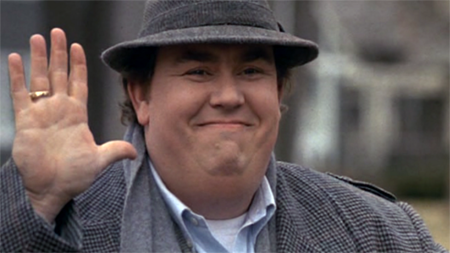 John Candy with his hand raised and wearing a fedora.