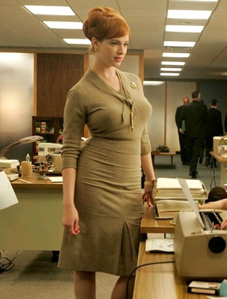 Christina in  navy green dress standing in front of a desk on the series 'Mad Men'.