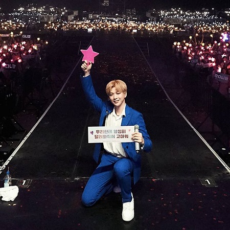 Kang holding a pink star in his right hand while kneeling in his blue dress at a stage's runway.