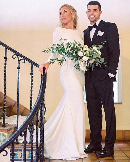 Allie and Jose in their wedding dresses standing in the staircase. Allie looking to her right holding a white flower bouquet and Jose hands in his pocket. 