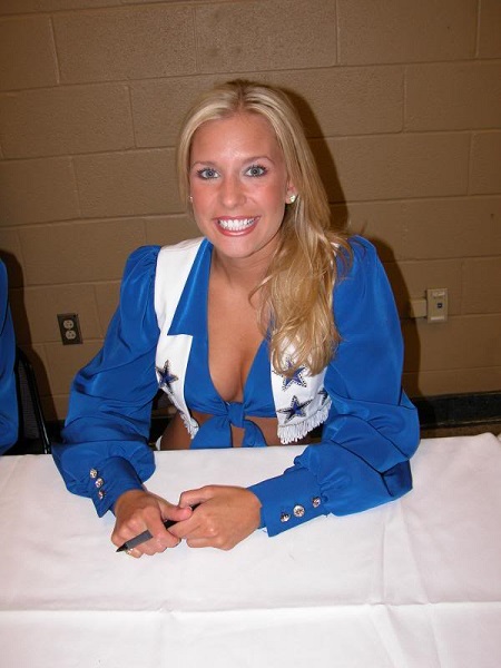 Emily sitting behind the desk in a blue cheerleading costume and smiling at the camera with a pen in her hand.