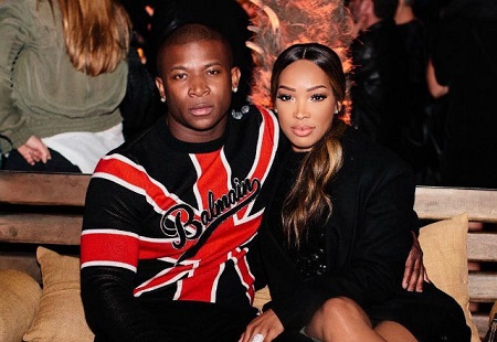 OT Genasis with his left arm around Malika as they face the camera sitting on a bench.