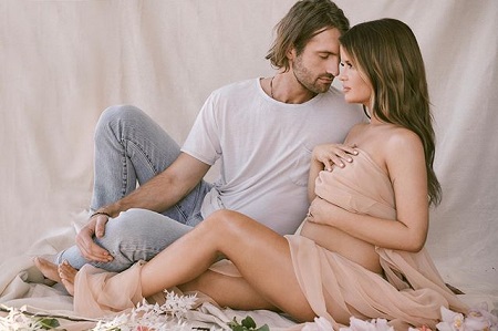 Maren Morris sitting on the floor touching her belly as Ryan closes in for a romantic pose with white background.