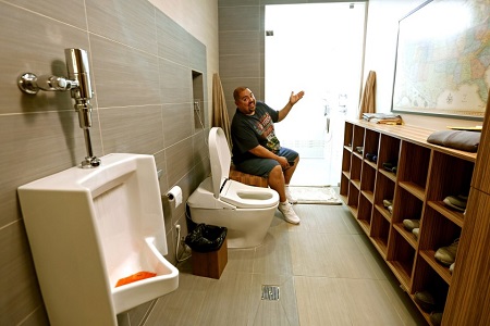 The bathroom is like a hallway with a shoe rack and toilet opposite to it. Fluffy is sitting on a seat next to it with his left arm raised up.