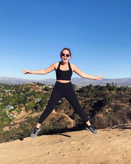 Burr jumping with her arms and legs spread in her black jogging suit at the edge of a cliff.