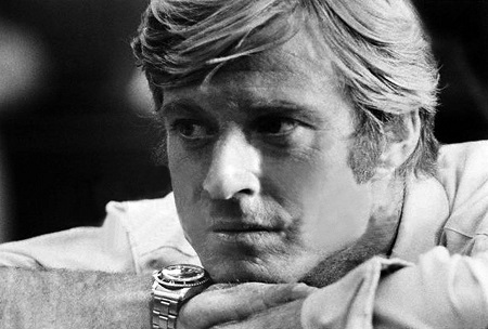 Black & White photo. Robert Redford in his younger years resting his chin in his hands sporting a watch in  his right hand.