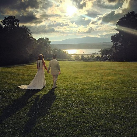 Two people (Molly and Ben) walking hand-in-hand on a green field overlooking a lake towards the eveining sunset.