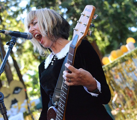 Kim Shattuck in front of a mic singing with a scream playing the guitar in a black dress.