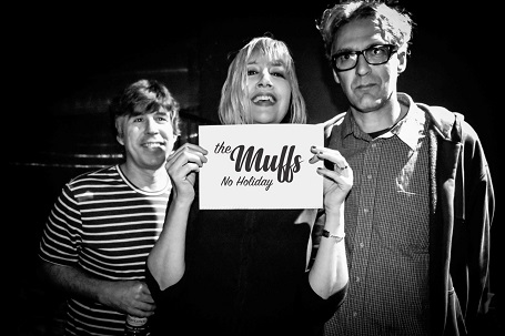 Shattuck holding a card that says 'The Muffs No Holiday' in the middle, with her band members on her sides.