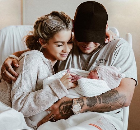 Kane and Katelyn on a hospital bed, Kane holding the baby in his arm with the other one around Katelyn whose hand is cusping the baby's left hand.