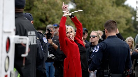 Jane Fonda raising her plastic-cuffed hands in the air with a crazy face, tongue sticking out.