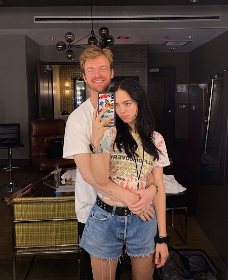 Claudia taking a mirror selfie with Finneas holding her from the back and smiling at the camera/mirror.
