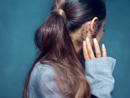 Ariana turning her head to the left pointing to the three tattoos on her right ear.