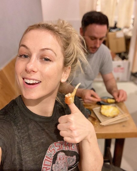 Iliza taking a selfie with her thumb wrapped around by food and Noah on the background.