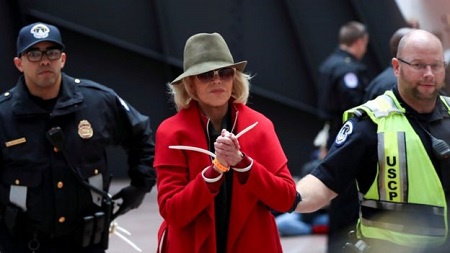 Jane Fonda wearing a red overcoat, a green hat and sunglasses while being arrested with two police authority people with her.