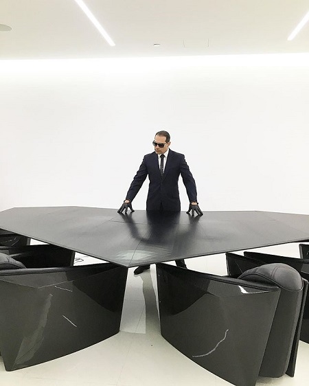 Manny Khoshbin posing in an all black stylish office table like James Bond in an all-white background.