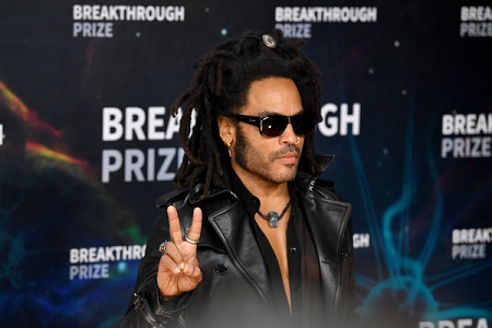 Lenny Kravitz in his black leather dress and glasses raising a peace sign with his right hand.