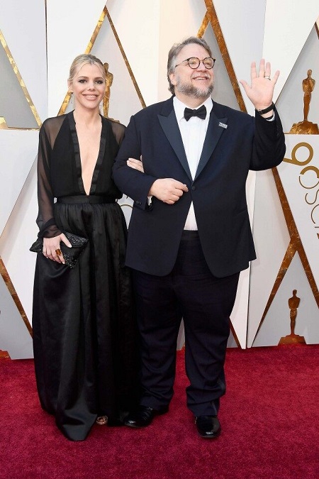 Kim Morgan in black dress (left) arm-in-arm with Guillermo del Toro (right)  waving her arm in the air. Both smiling.