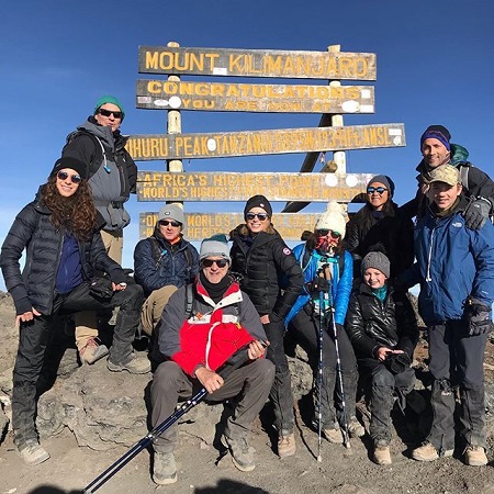 Amy and Andrew in front of the congratulations board on the summit of Mount Kilimanjaro with all their close friends wearing buffy clothes.