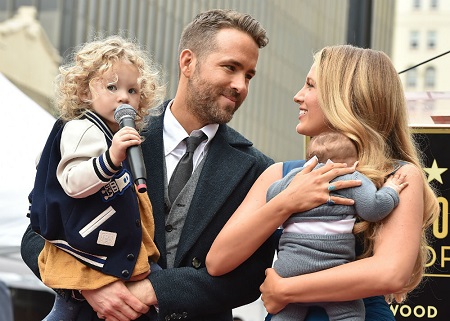 Ryan Reynolds holding older daughter James in his arms as she holds a mic to her face, and his wife, Blake Lively holding the still-toddler second daughter, Inez, in her arms.