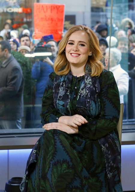 Adele sitting on a chair smiling at the camera. Glass window on the background with paparazzis.