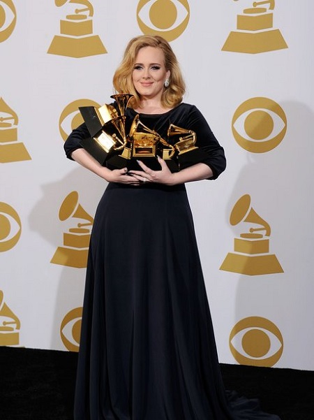 Adele in black holding six Grammys in her arms smiling at the camera.