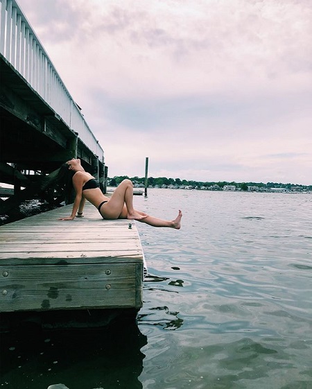 Charli D'Amelio in her black swimsuit from a far and side angle kicking back the water while sitting in the docks.