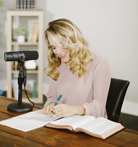 Jinger Duggar in front of a table microphone for her podcast writing something down on paper with a bible beside her.